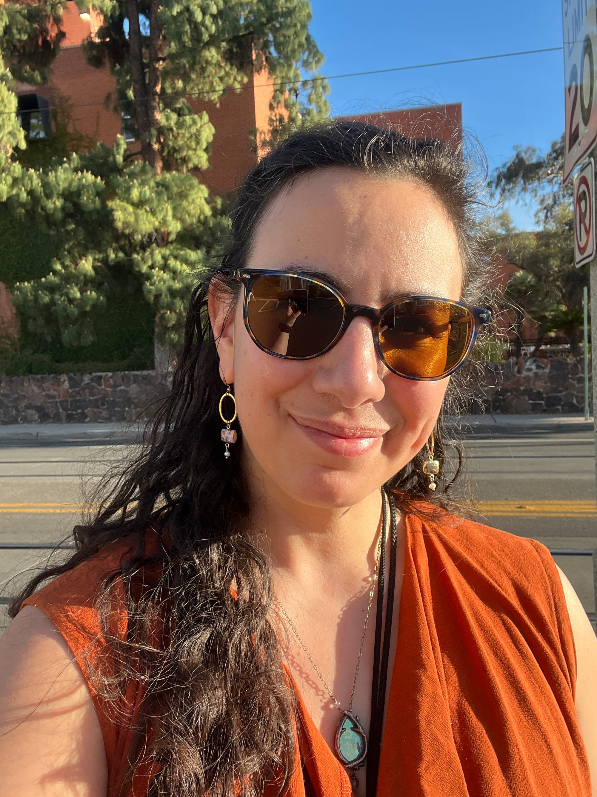 A sunny selfie in which I’m wearing roundish sunglasses, my long dark wavy hair half up, half down, an orange sleeveless cowl-necked top, as well as earrings that dangle a black pearl rectangle and white pearl bead beneath a gold hoop, and a Parrish Relics necklace that encloses a floral tapestry image under reflective mica between two brass branches that bend to make an oval together.