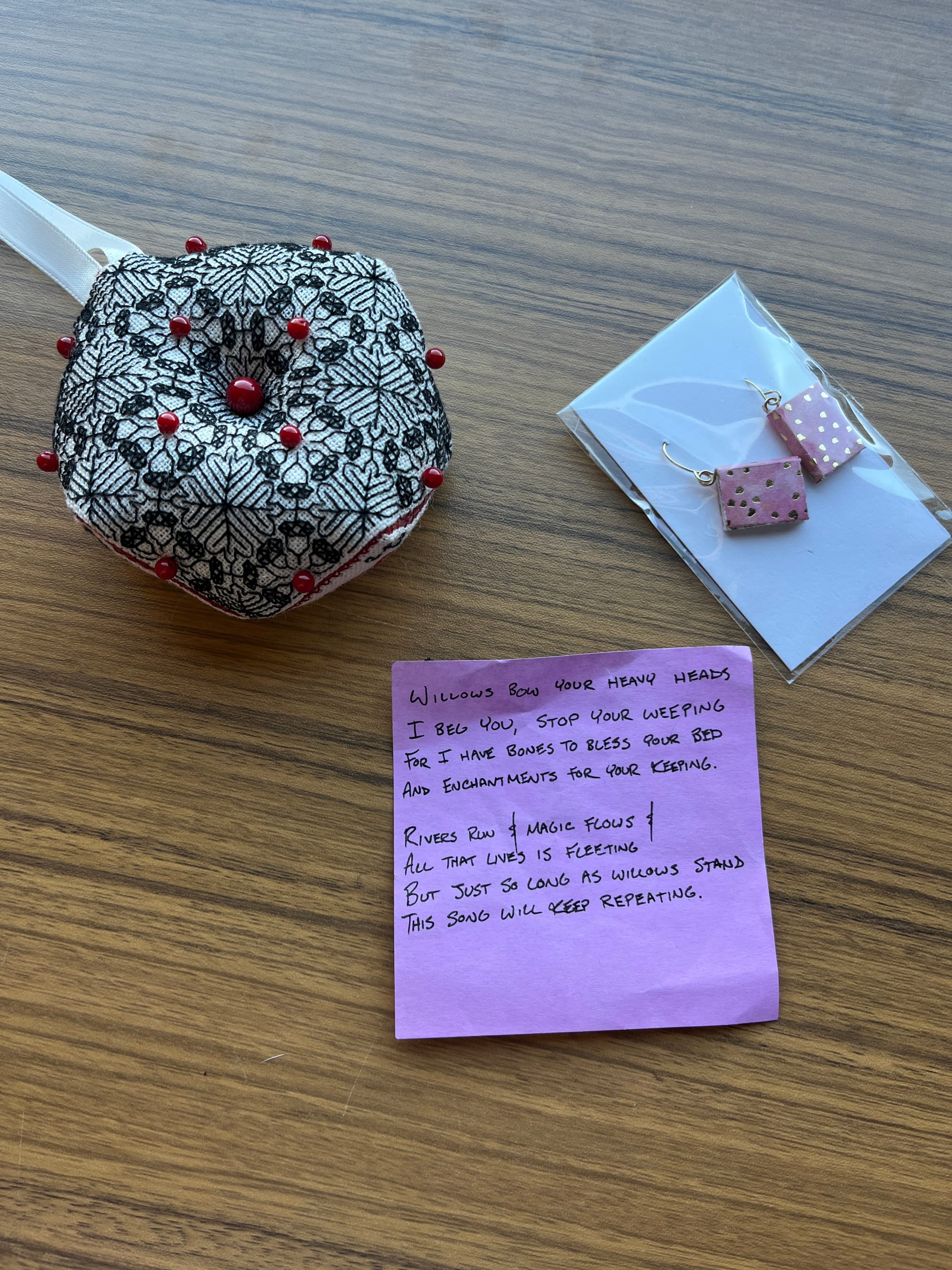 A photo of an embroidered ornament, showing a pattern of acorns and oak leaves in a geometric design, dotted with red beads. To the right of it is a pair of pink book-shaped earrings with gold flecks on the covers; below them is a purple post-it note on which is written the following poem. "Willows bow your heavy heads / I beg you, stop your weeping / For I have bones to bless your bed / and enchantments for your keeping / / Rivers run and magic flows / all that lives is fleeting / but just so long as willows stand / this song will keep repeating."