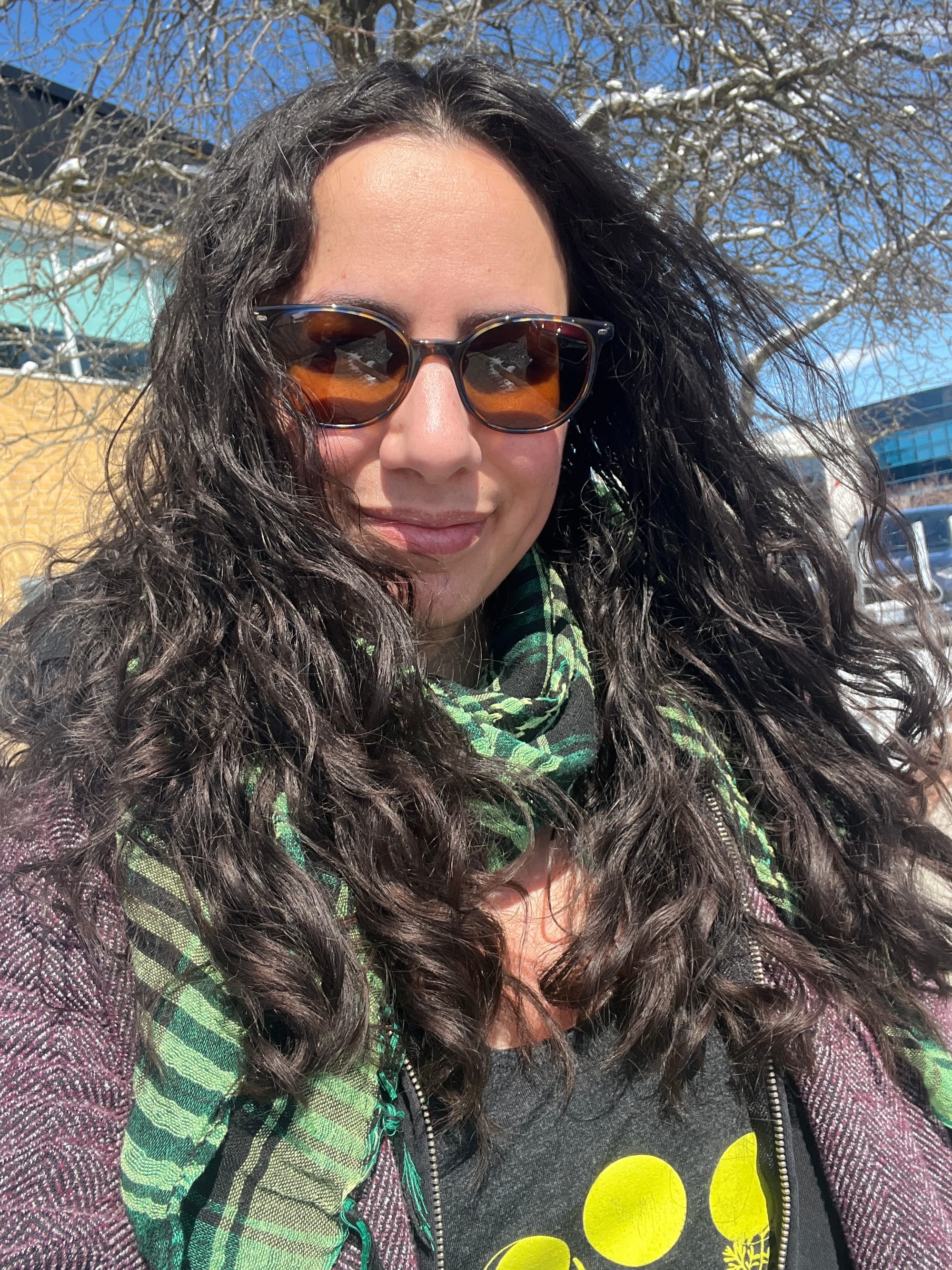 Sunny selfie in which my long dark hair is worn down and looks frankly amazing in a tumult of wavy curls falling over my shoulders. I'm wearing sunglasses, a green and black keffiyeh, a black tank top with bright yellow circles visible on it under a black hoodie under a dark purple and black wool jacket woven in a chevron pattern. Behind me are snowy bare branches, buildings, and a bright blue sky.