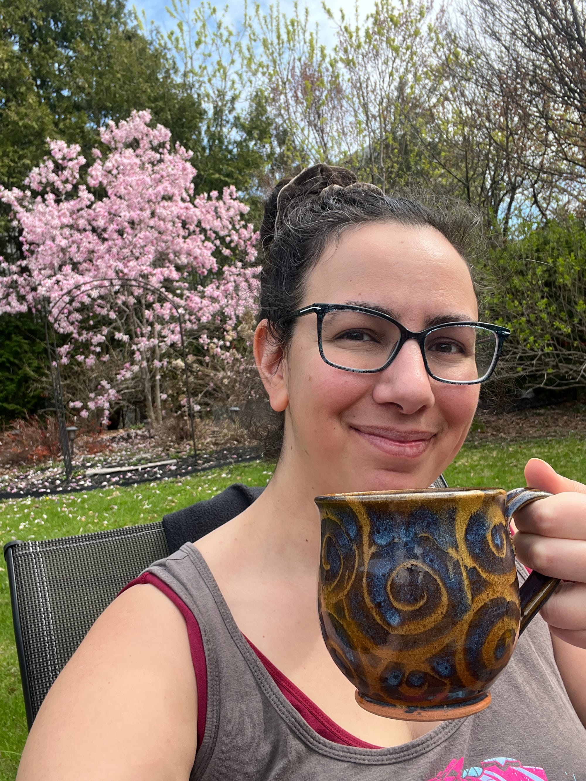 Selfie in which I'm sitting in a garden with a bright pink magnolia tree in the background, while holding up a big mug of tea and smiling. 