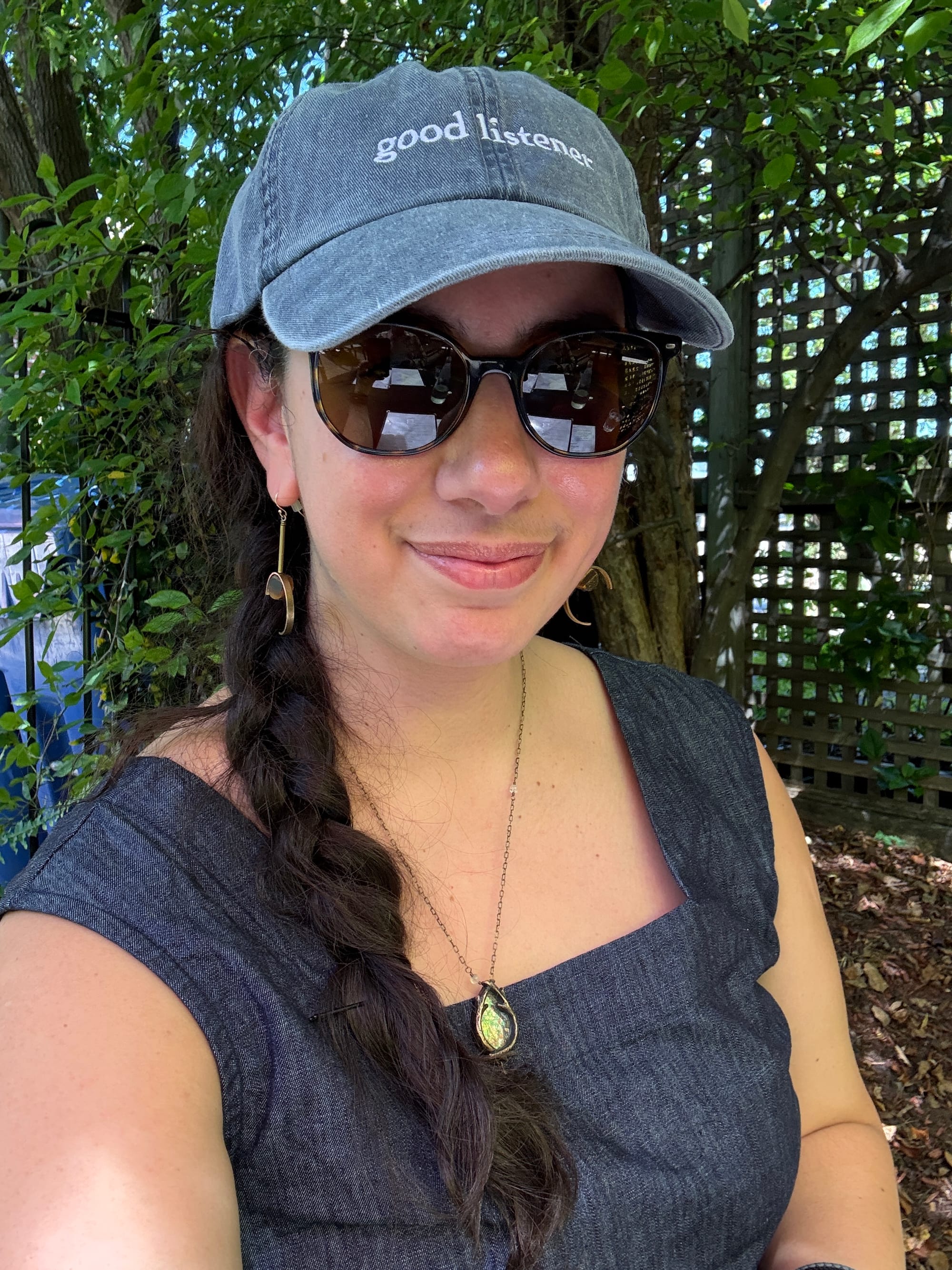 A selfie taken against a background of leafy green trees and a square trellis. I'm smiling close-mouthed while wearing roundish sunglasses, a dark blue sleeveless linen top, my long dark hair in a side braid, and a pale denim baseball cap from Macmillan Audio that reads "Good Listener" on the front. I'm also wearing dangly earrings with a brassy half-moon shape at the end inside of which is nestled a round flat wooden bead, as well as a Parrish Relics necklace that consists of a pair of brass branches enclosing a piece of William Morris foliate wallpaper.