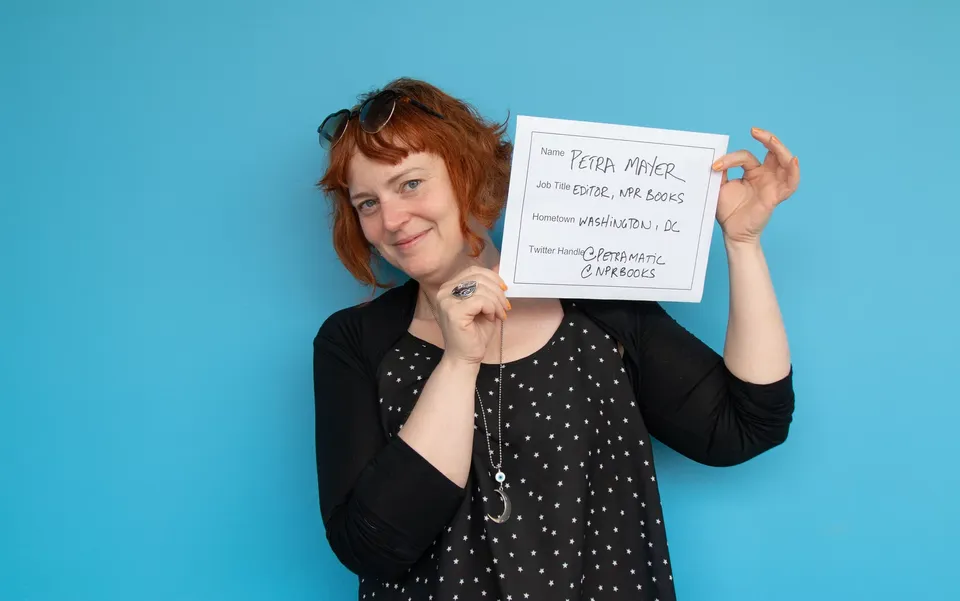 Petra Mayer smiling against a light blue background, holding a paper with her name, job, hometown and twitter handles on it.