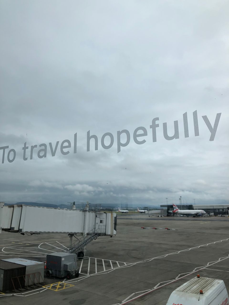 A grey day over Glasgow airport's airfield, glimpsed through a window on which the words "To travel hopefully" appear.