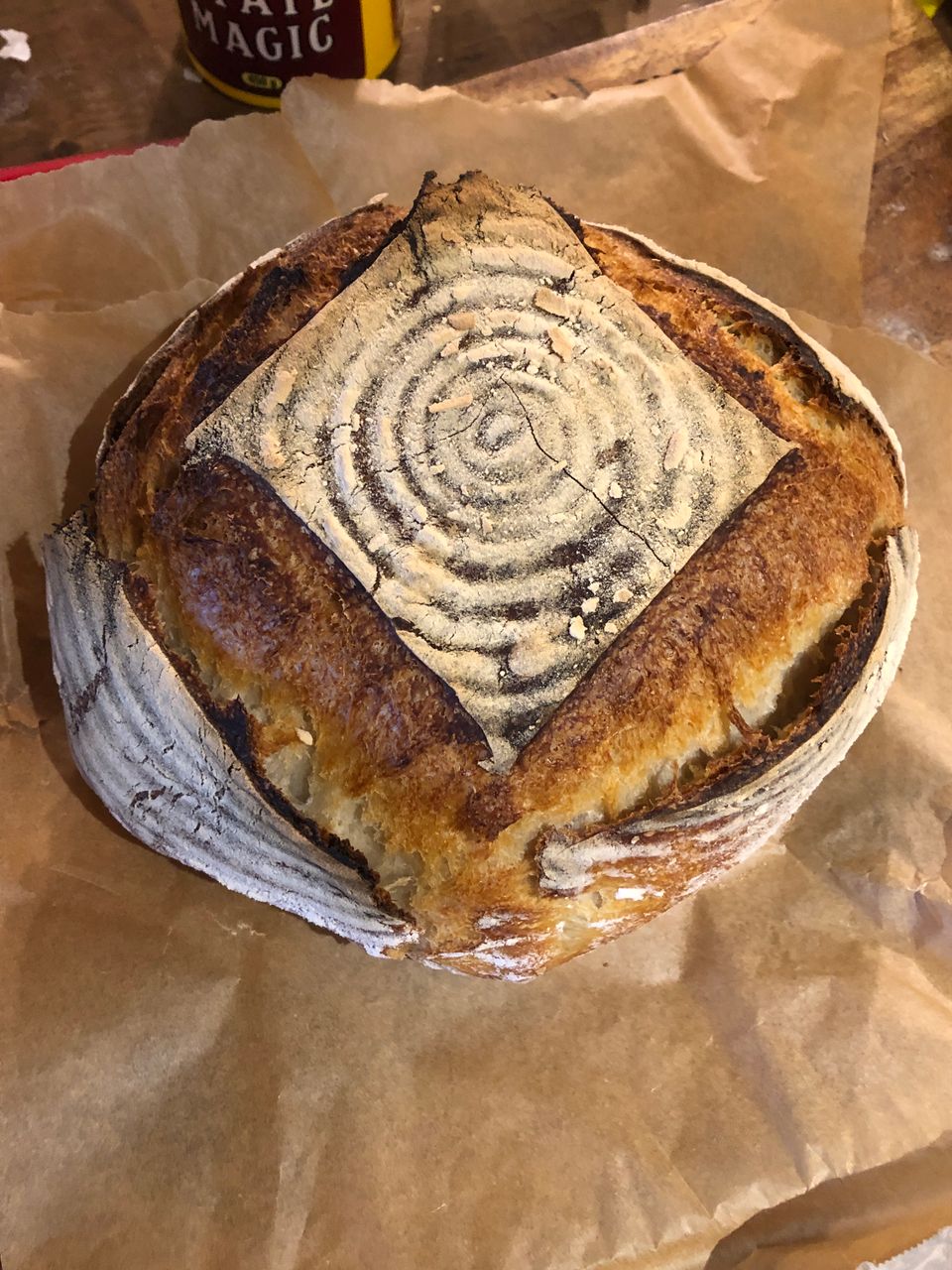 A fresh-baked sourdough boule, slashed such that a square of concentric flour circles sits decoratively atop the loaf. 