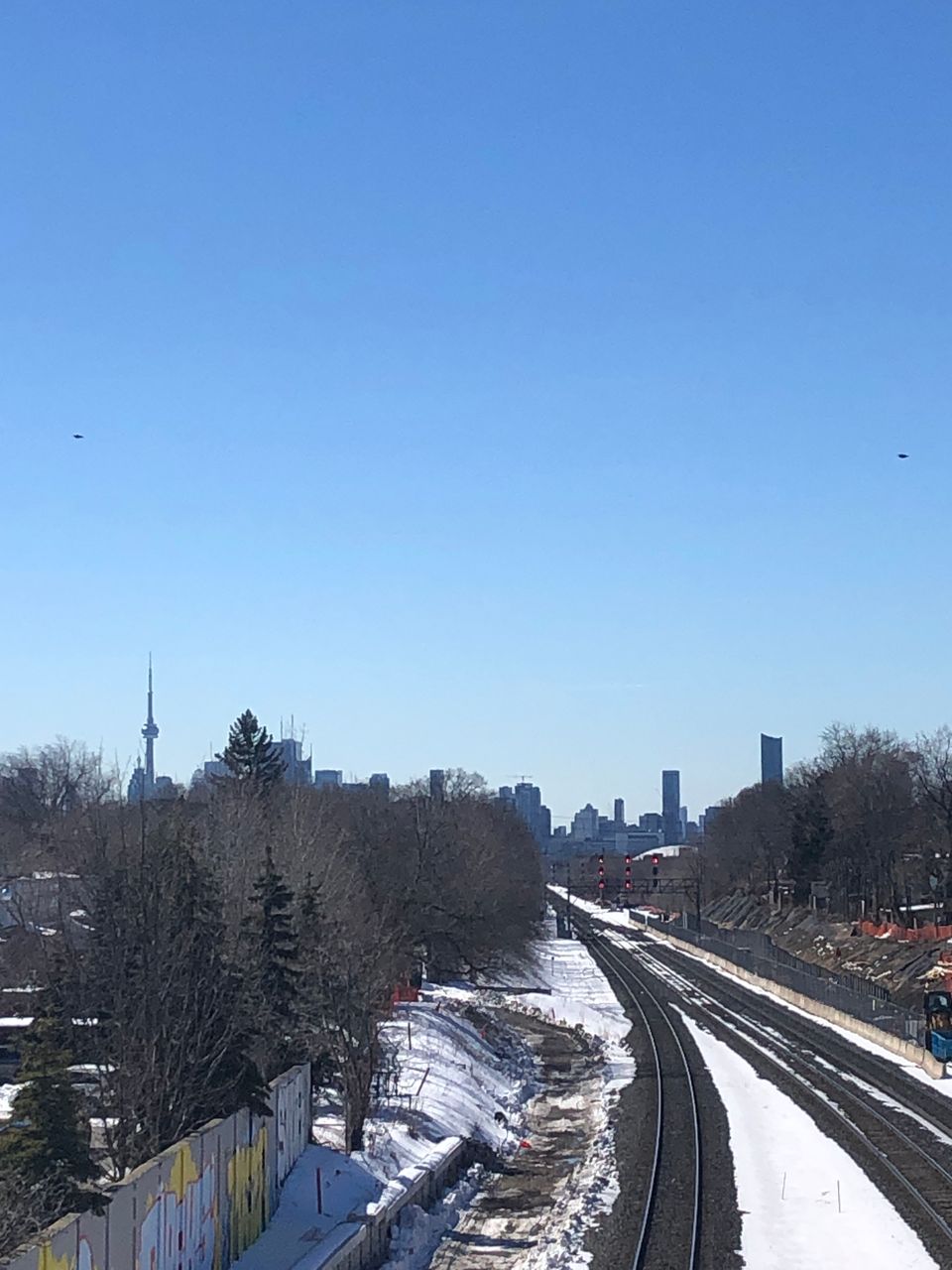 Snowy train tracks flanked by trees under a blue sky, with the city of Toronto silhouetted in the distance.